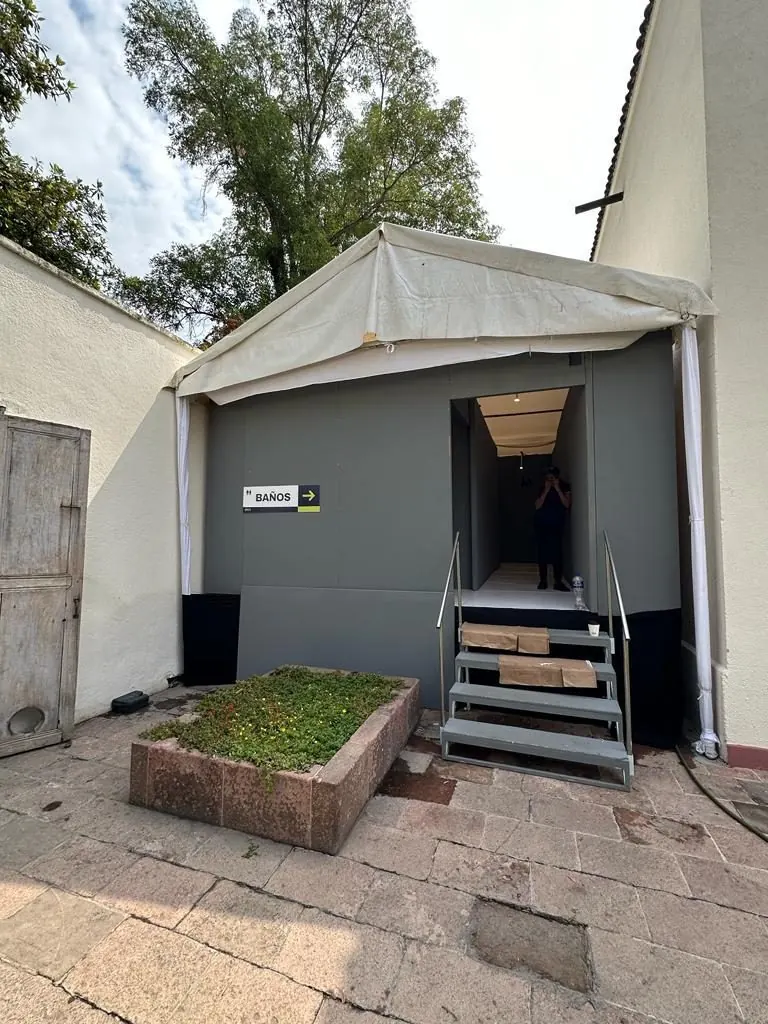 Módulo de baños de lujo VIP en gris bajo una carpa, con escalera de acceso, en un patio empedrado
