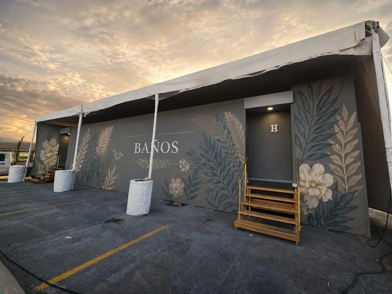 Fachada de unos baños portátiles para evento con mural de hojas y flores, escalera de madera y carpa, al atardecer