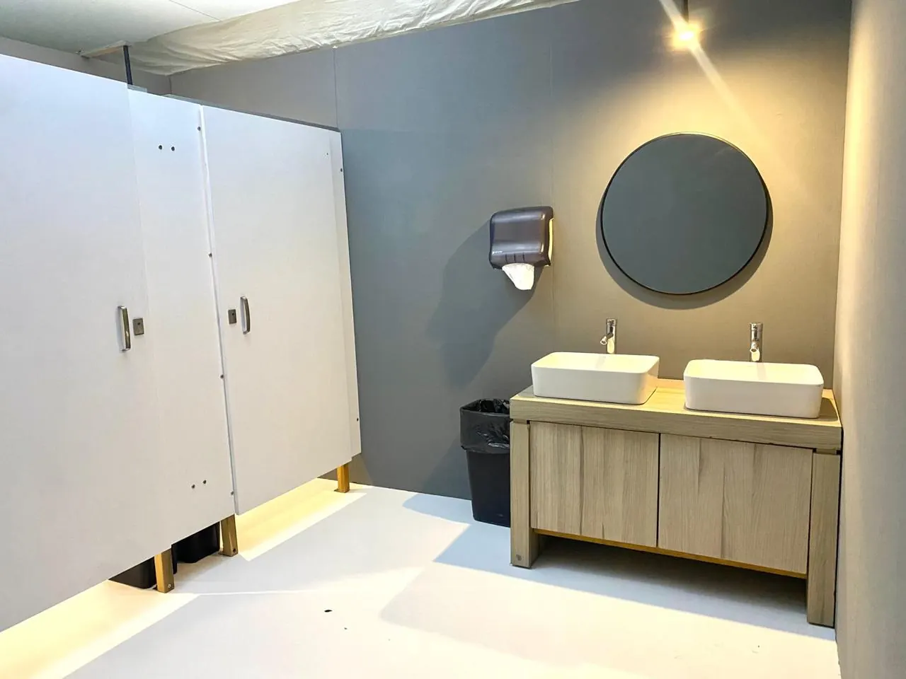 Interior de un baño portátil de lujo VIP en gris, con cubículos blancos, lavabo doble de madera y espejo redondo