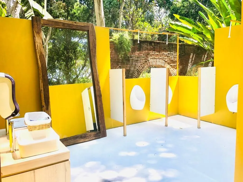 Baño de lujo al aire libre con muros amarillos, espejo de marco de madera antigua y mingitorios con divisores