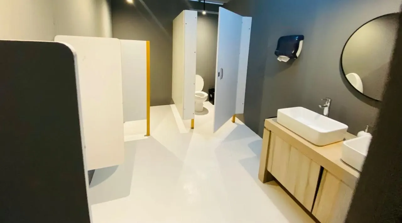 Pasillo de cubículos blancos en un baño portátil de lujo VIP gris, con lavabos sobre mueble de madera y espejo redondo