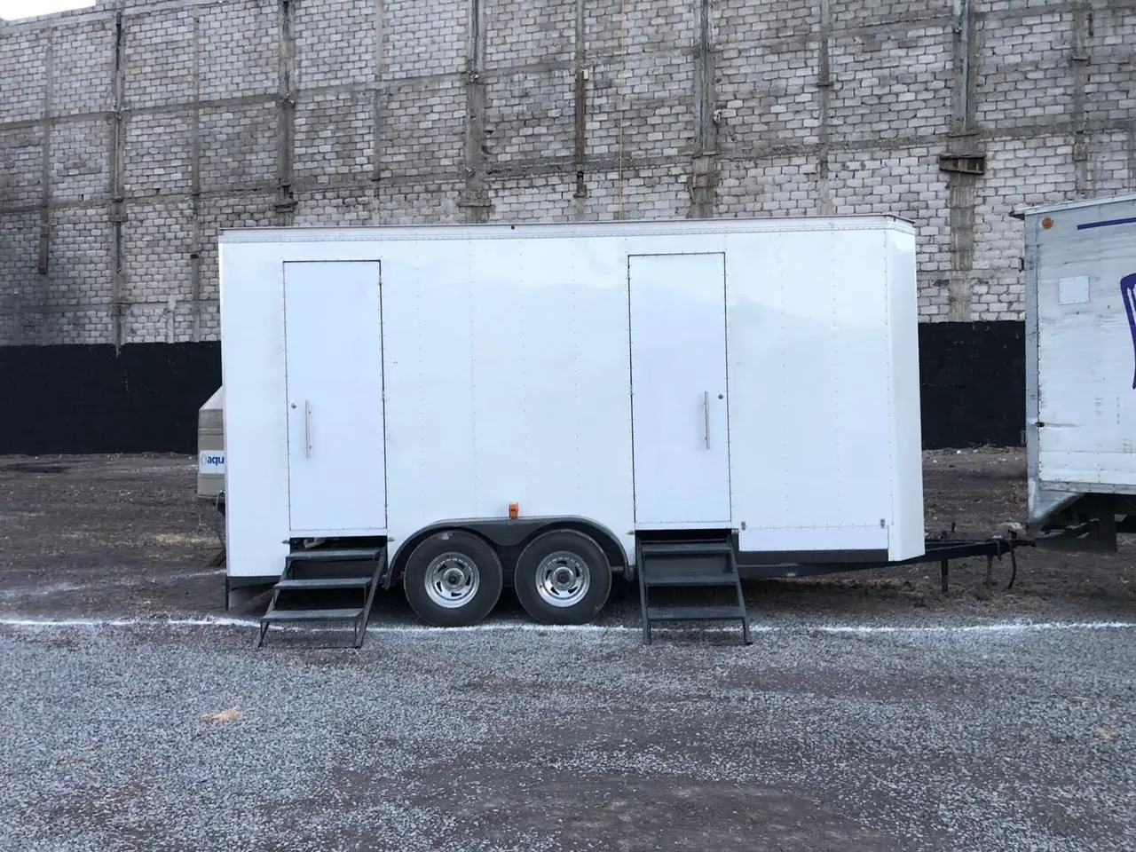 Baño remolque VIP blanco con dos puertas y escaleras metálicas, estacionado sobre grava