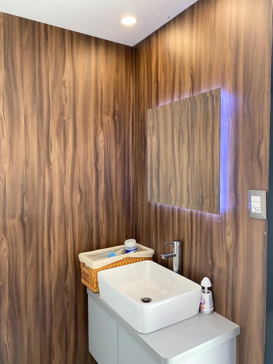 Lavabo blanco con llave de agua y espejo con luz LED dentro del baño remolque VIP, con paredes de madera