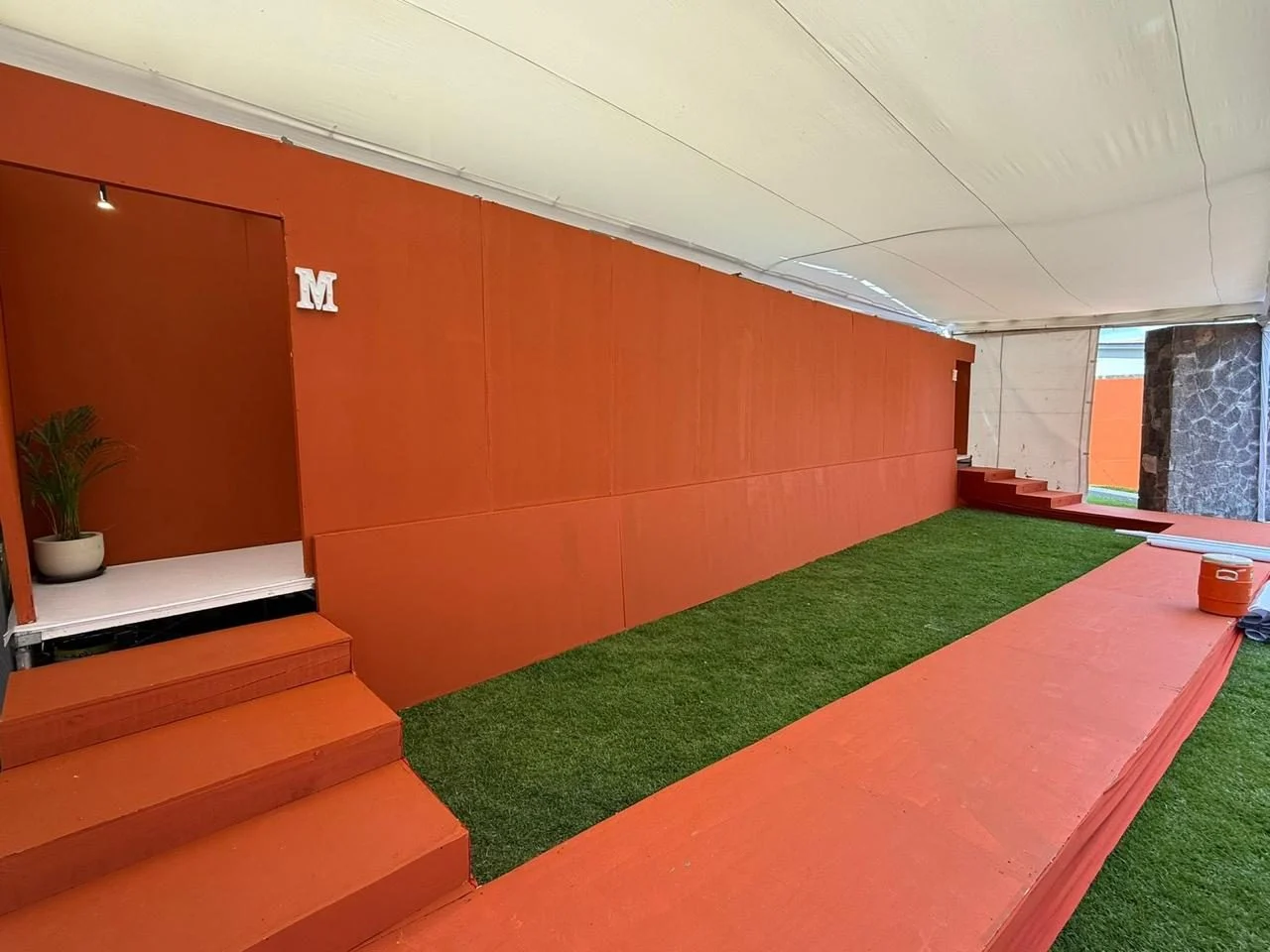 Entrada de un baño VIP montado bajo carpa, con muros naranjas, escalones y pasto
