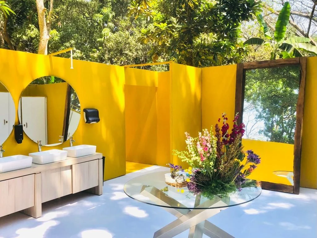 Baño VIP amarillo al aire libre con lavabos, espejos redondos y una mesa de cristal con flores
