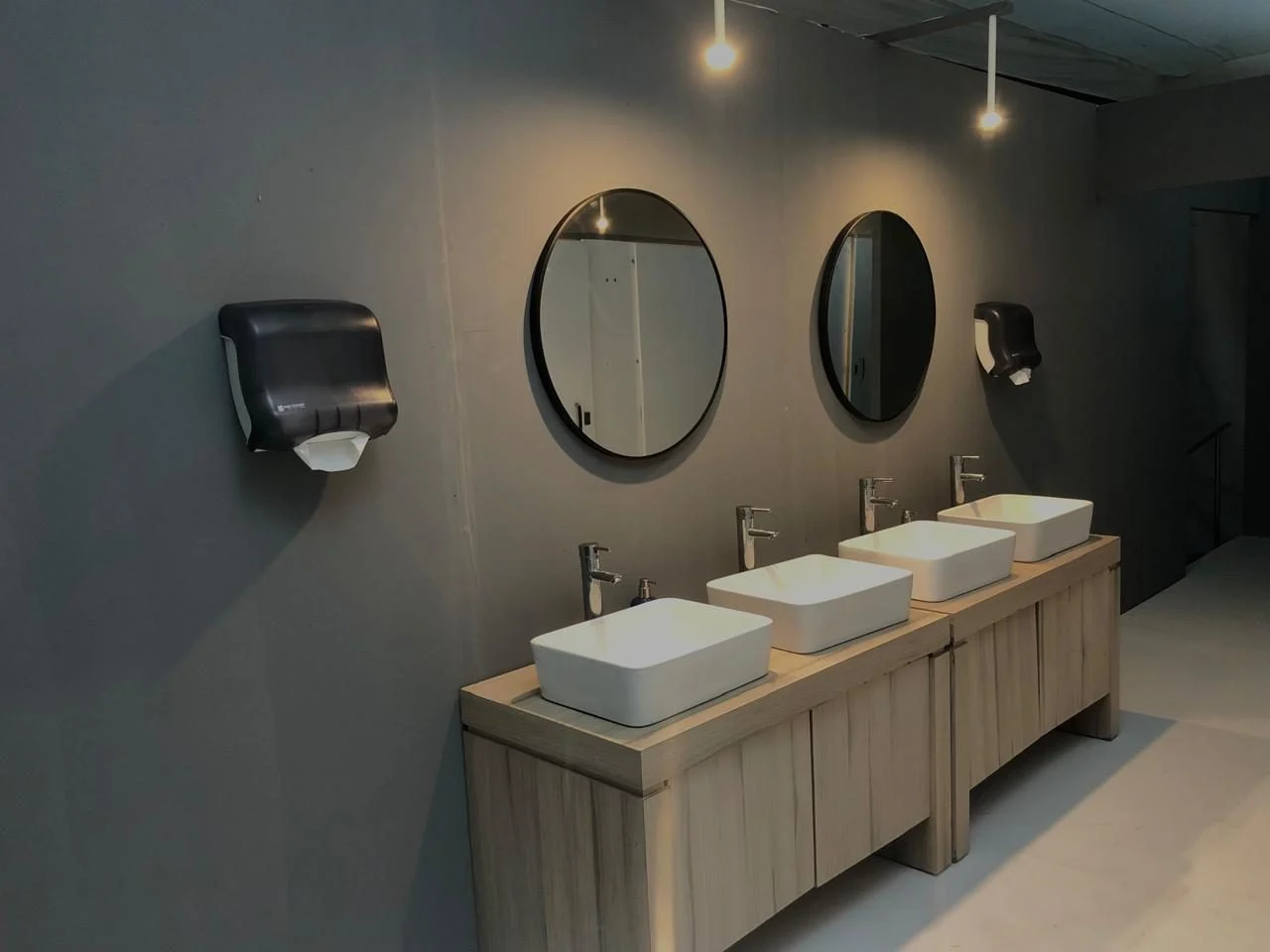 Tocador de madera con tres lavabos y espejos redondos dentro de un baño VIP de muros grises