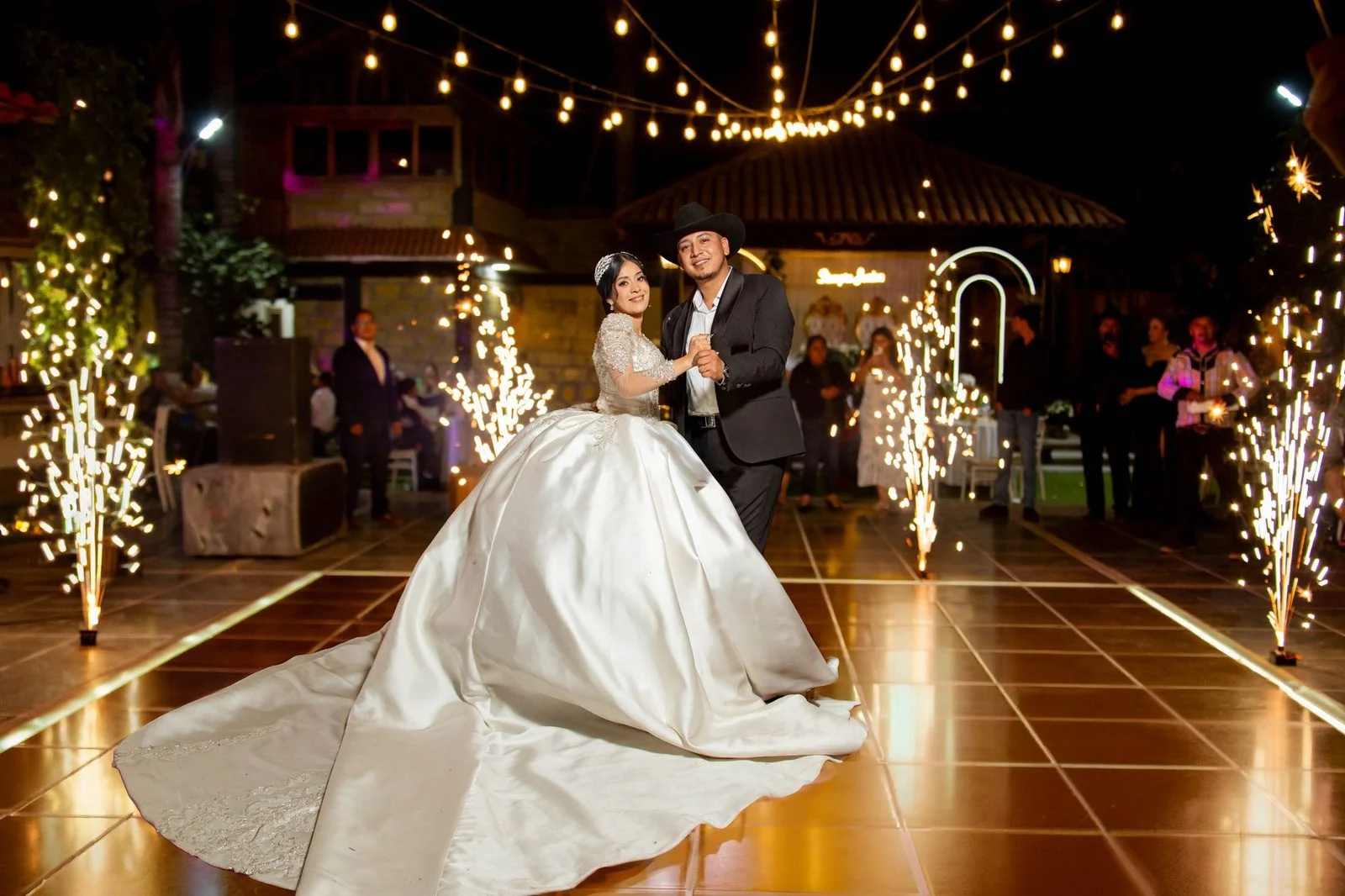 Novios bailando de noche en una pista con luces colgantes y chisperos, invitados alrededor